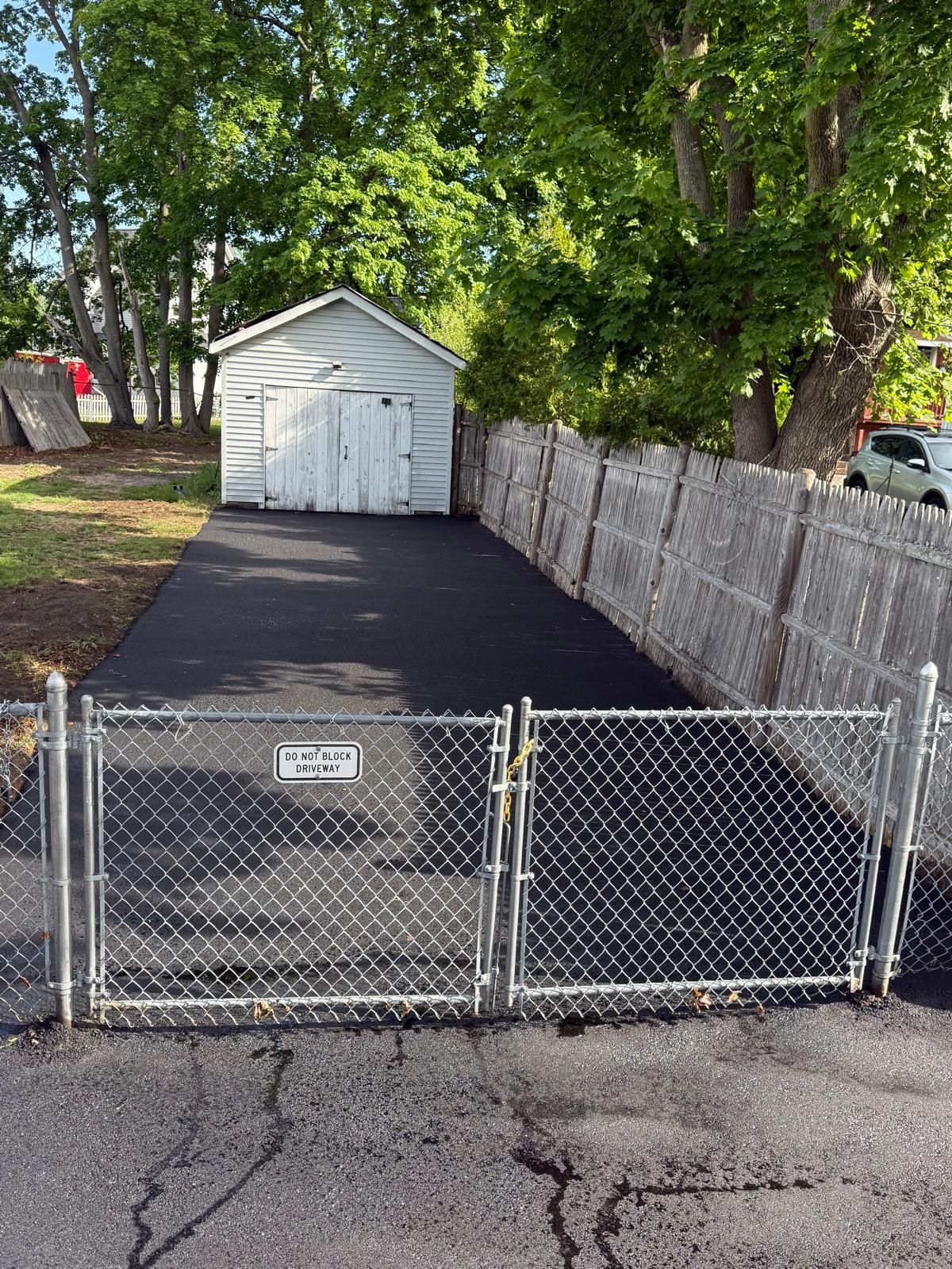 Chain-link fenced driveway with black asphalt leading to a white shed under trees