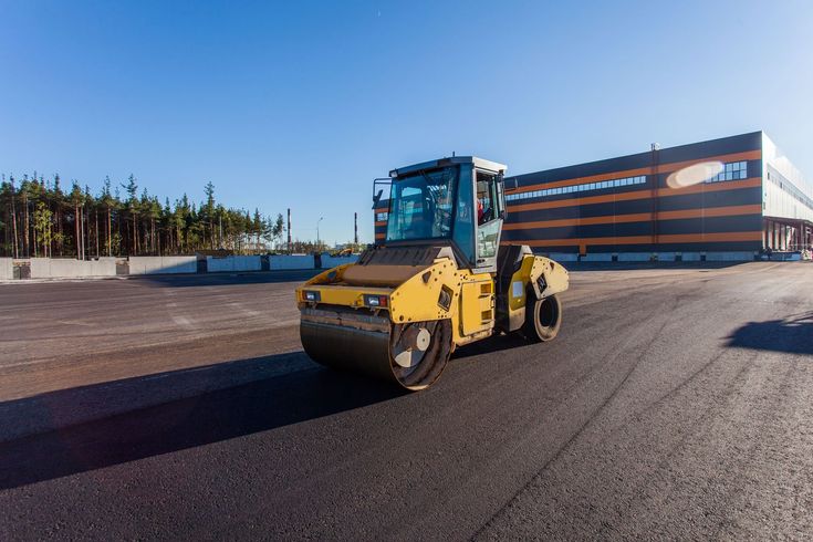 Yellow road roller on a paved surface near a striped building under a clear blue sky
