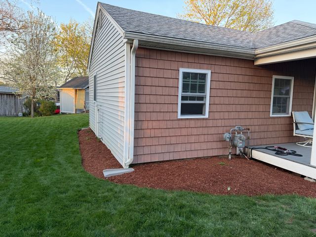 Backyard side of a house with tan siding, white trim, green lawn, and red mulch beds