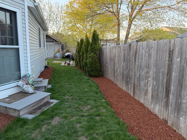 Narrow side yard with house steps, green grass, red mulch border, wooden fence, and shrubs.