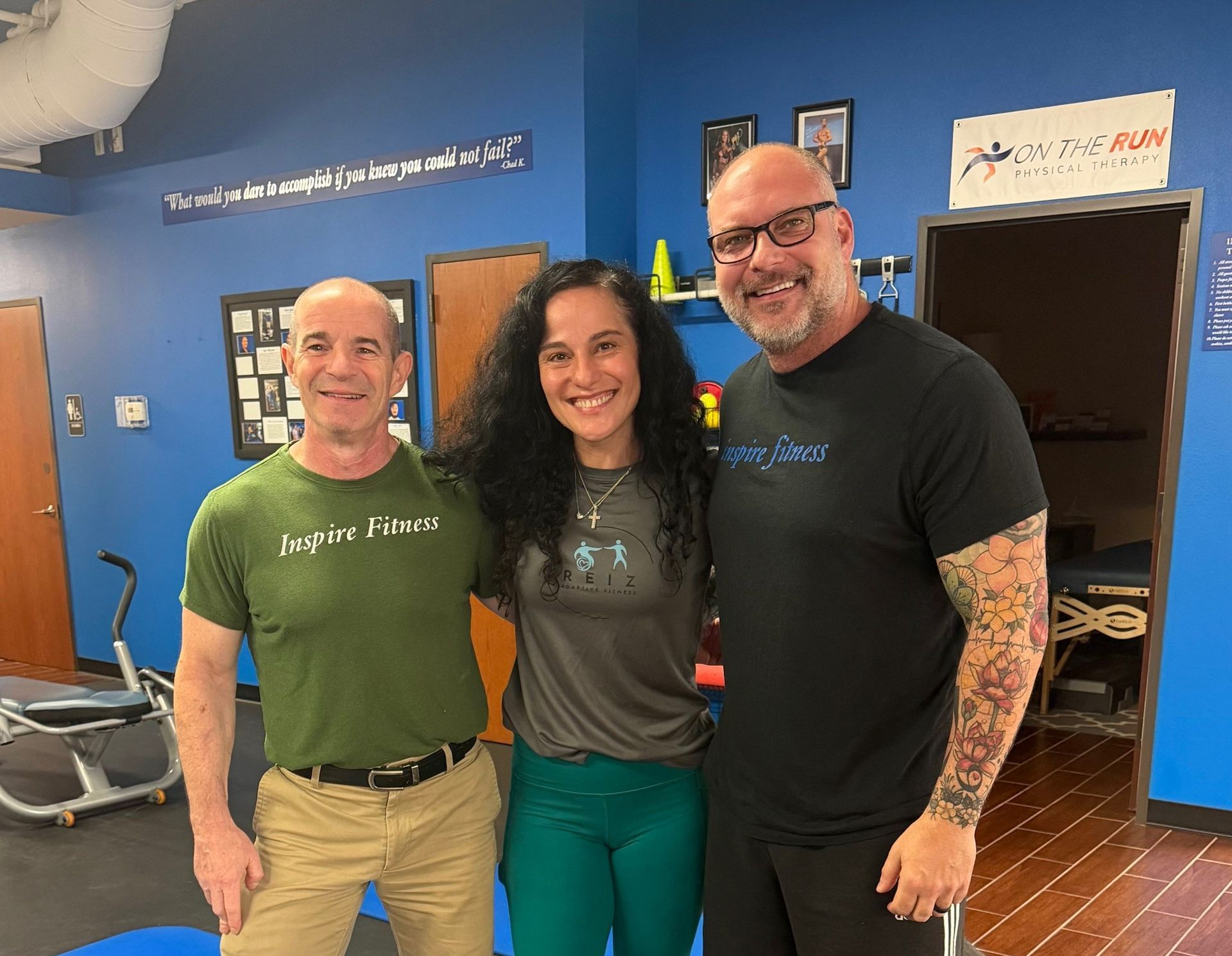 Three people standing together in a gym, smiling beside exercise equipment and blue walls.