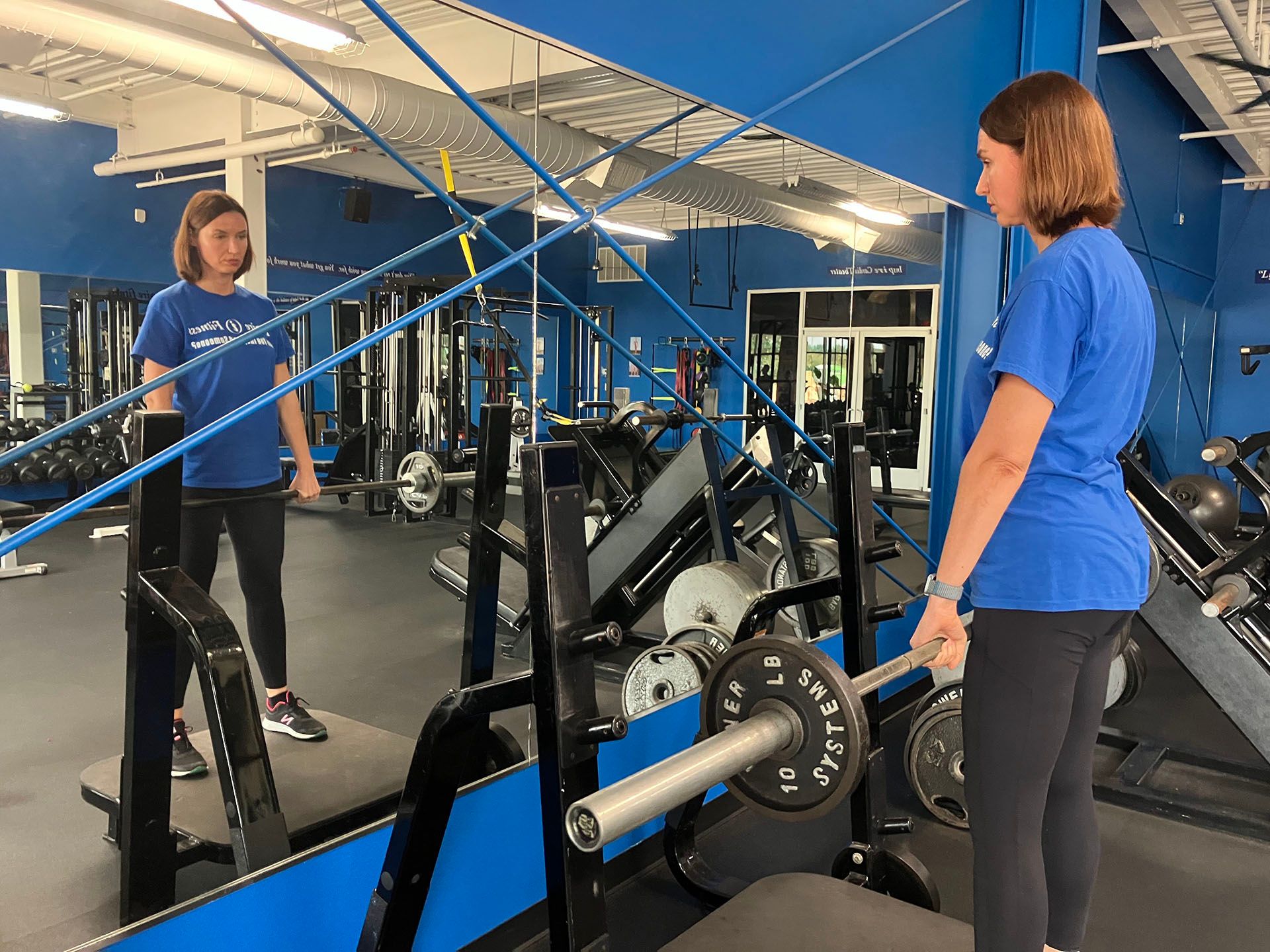 Woman in blue shirt standing beside a barbell in a mirrored gym.