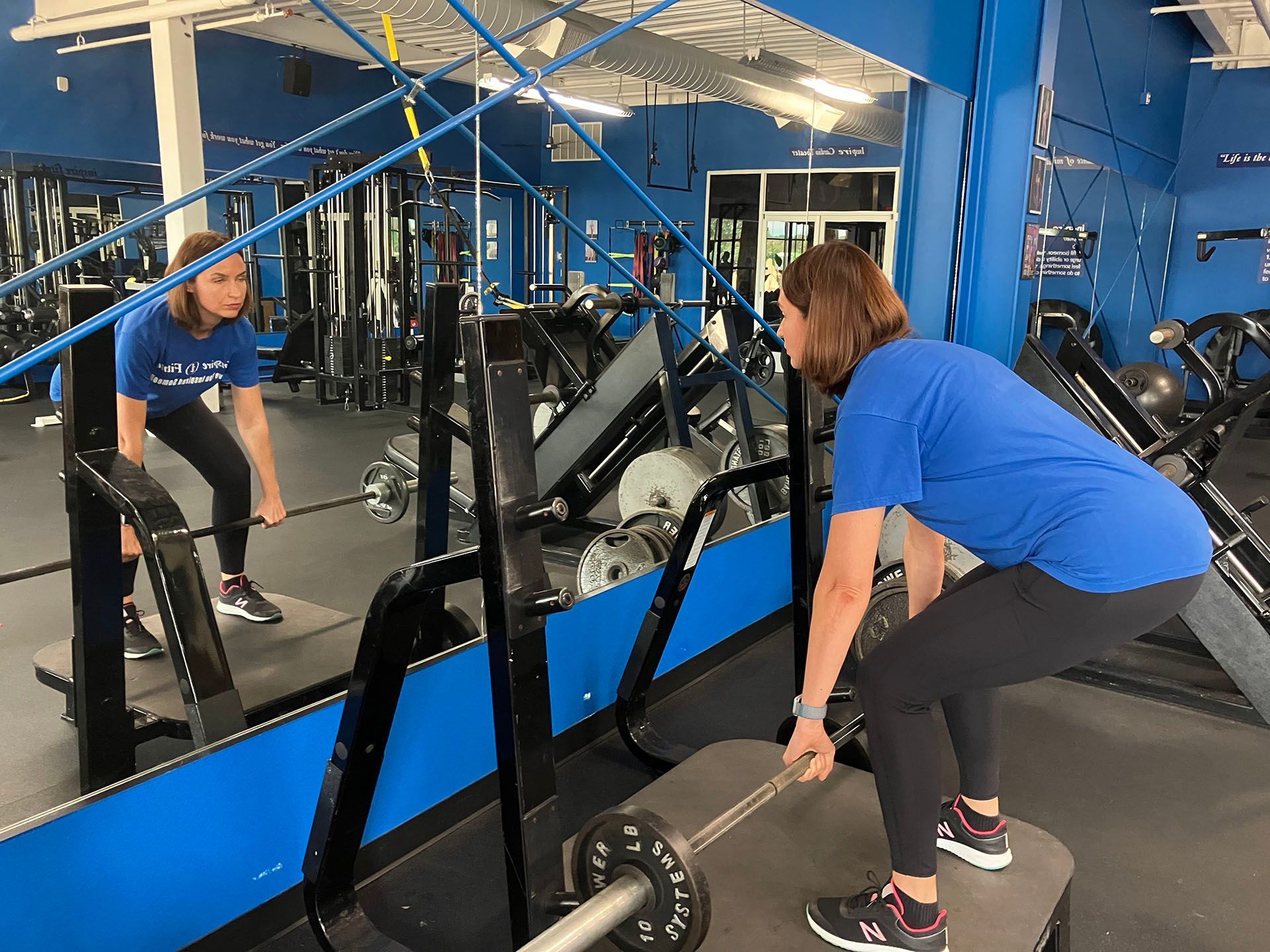 Woman lifting a barbell in a mirrored gym, wearing a blue shirt and black leggings.