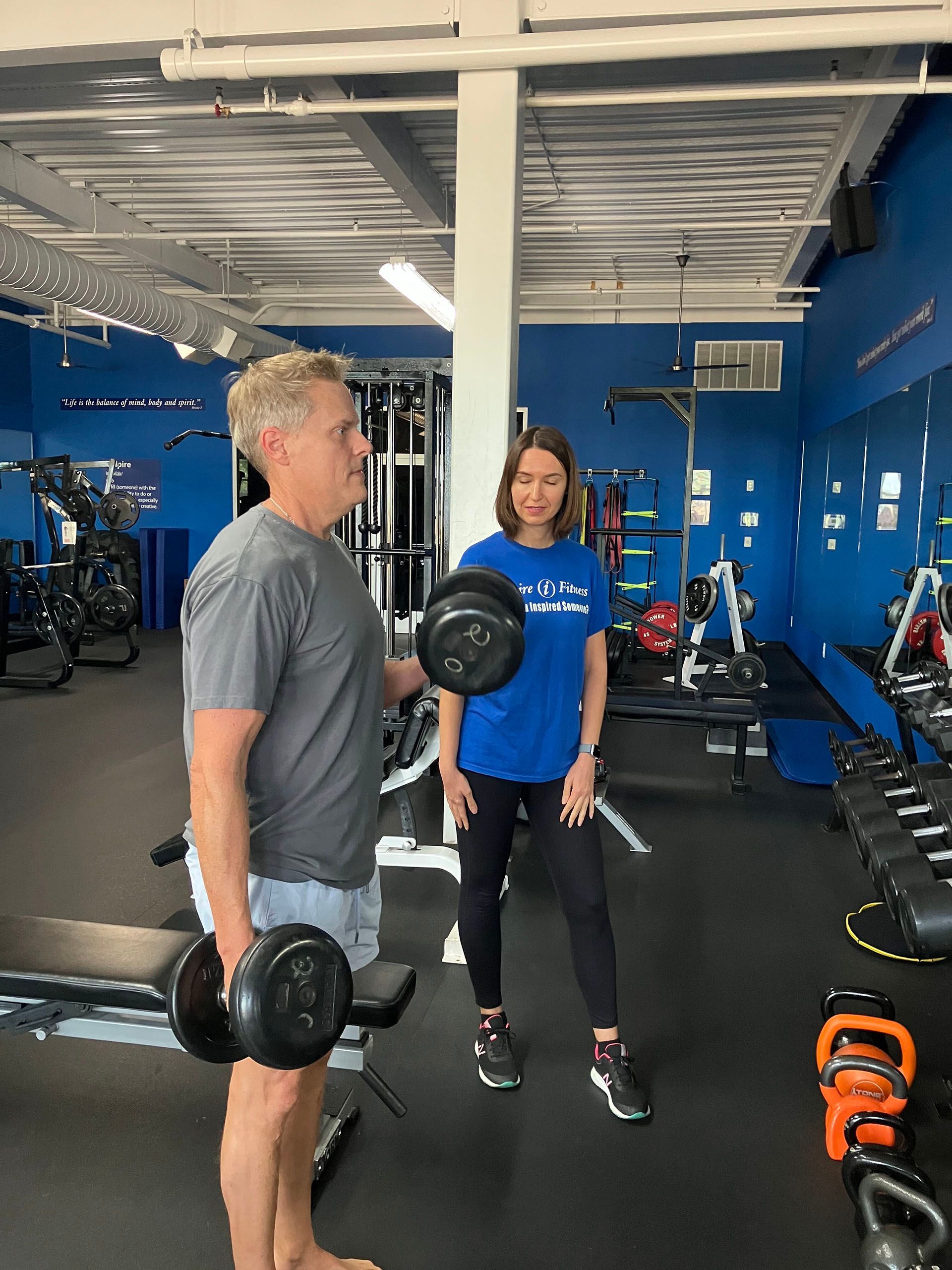 Two people holding dumbbells in a blue gym, standing near exercise equipment.