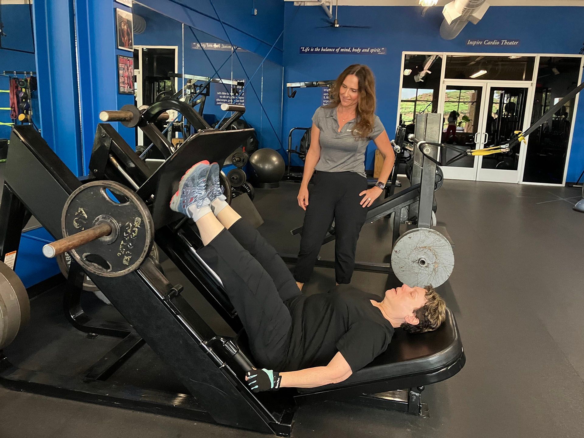 Person using a leg press machine in a gym while another person stands nearby.