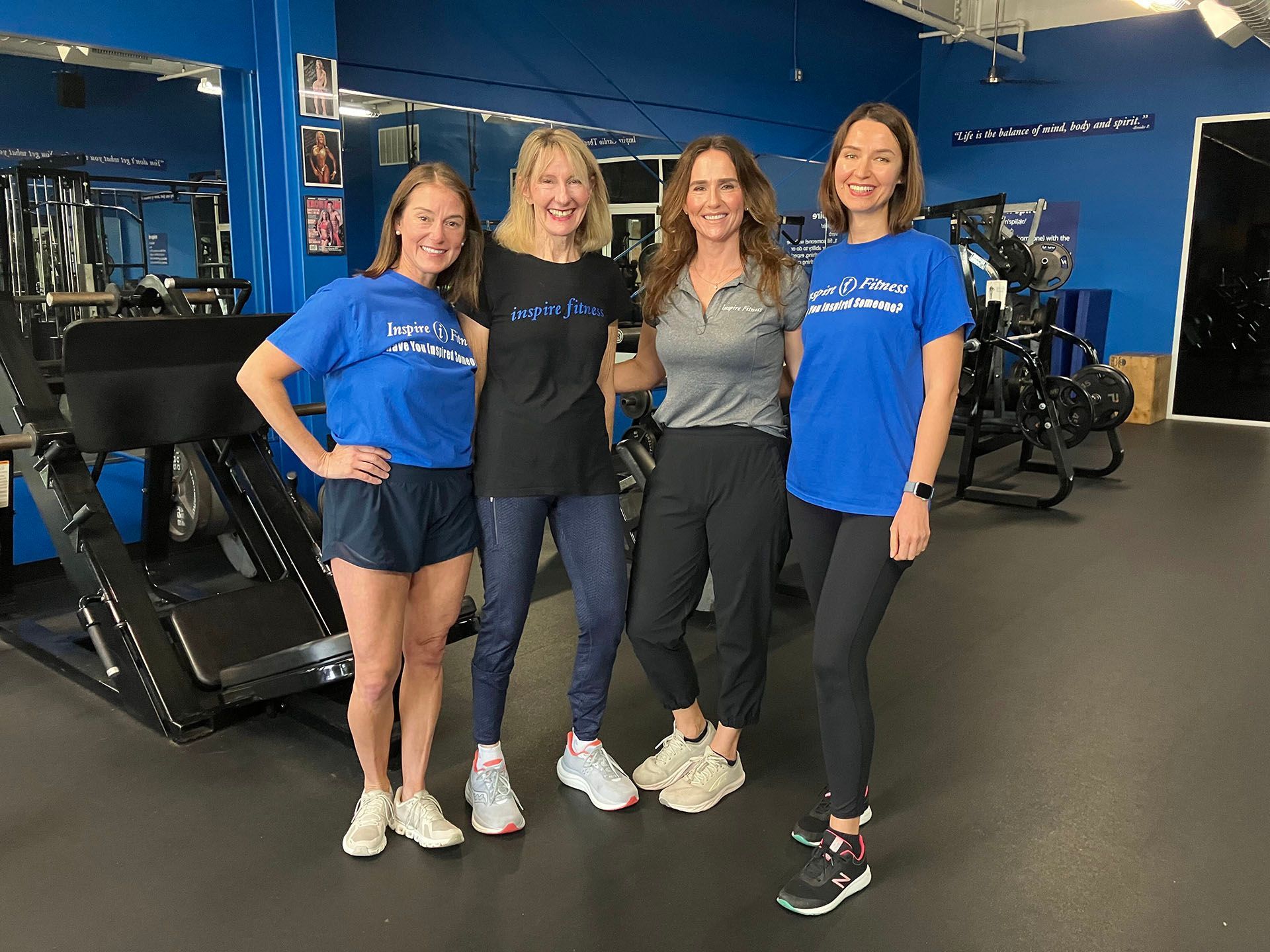 Four women posing together in a gym, smiling in front of exercise equipment.