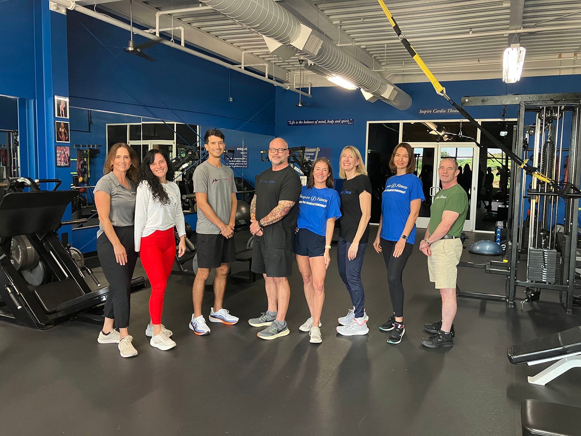 Group of seven people standing in a gym, posing for a photo near exercise equipment.