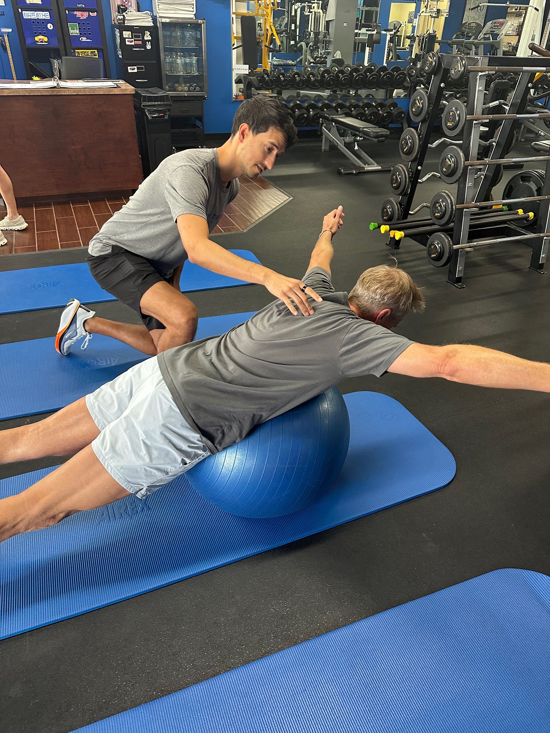 Trainer assisting person balancing on blue exercise ball in a gym.
