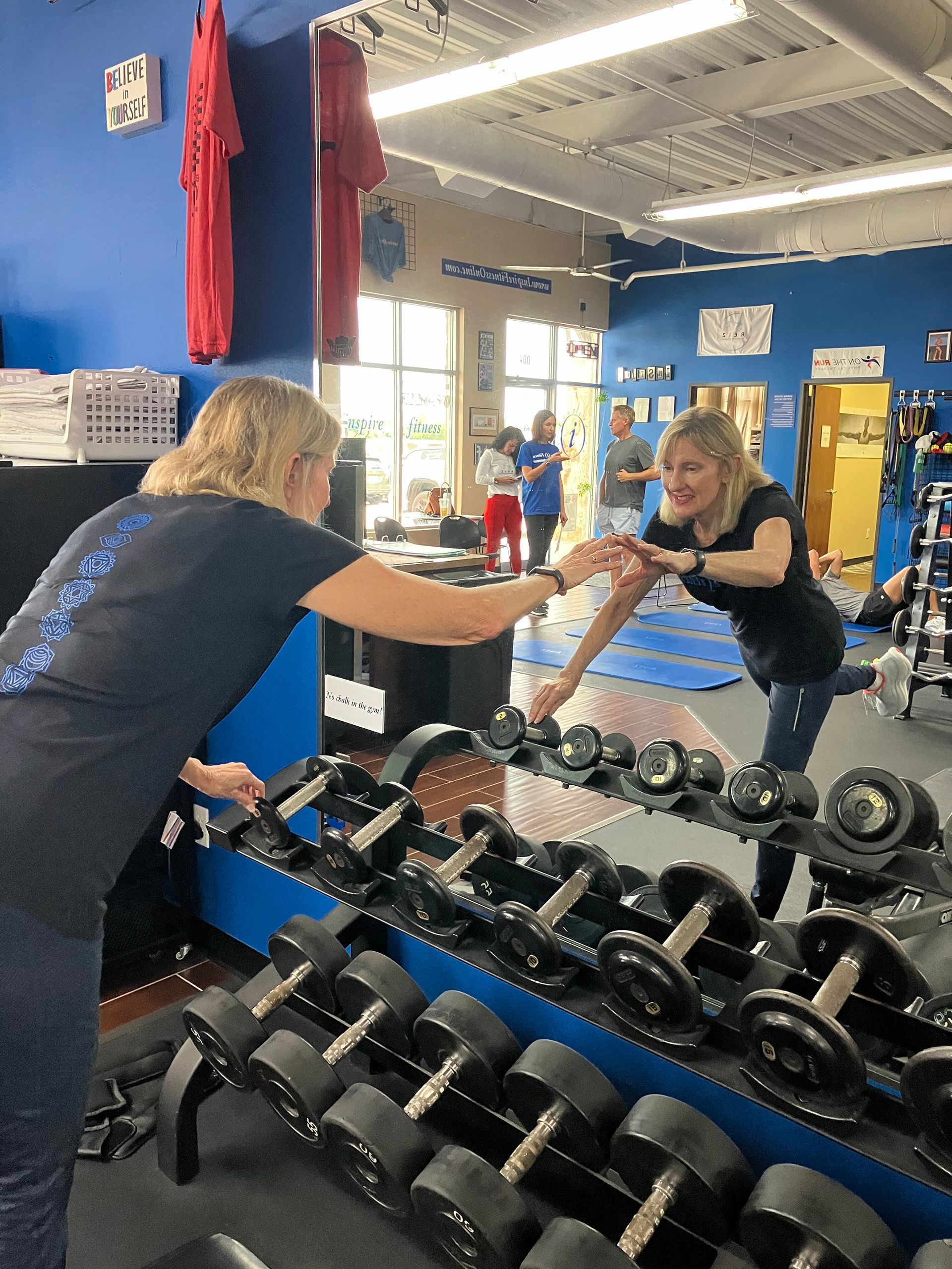 Two people in a gym do a high-five across a dumbbell rack.