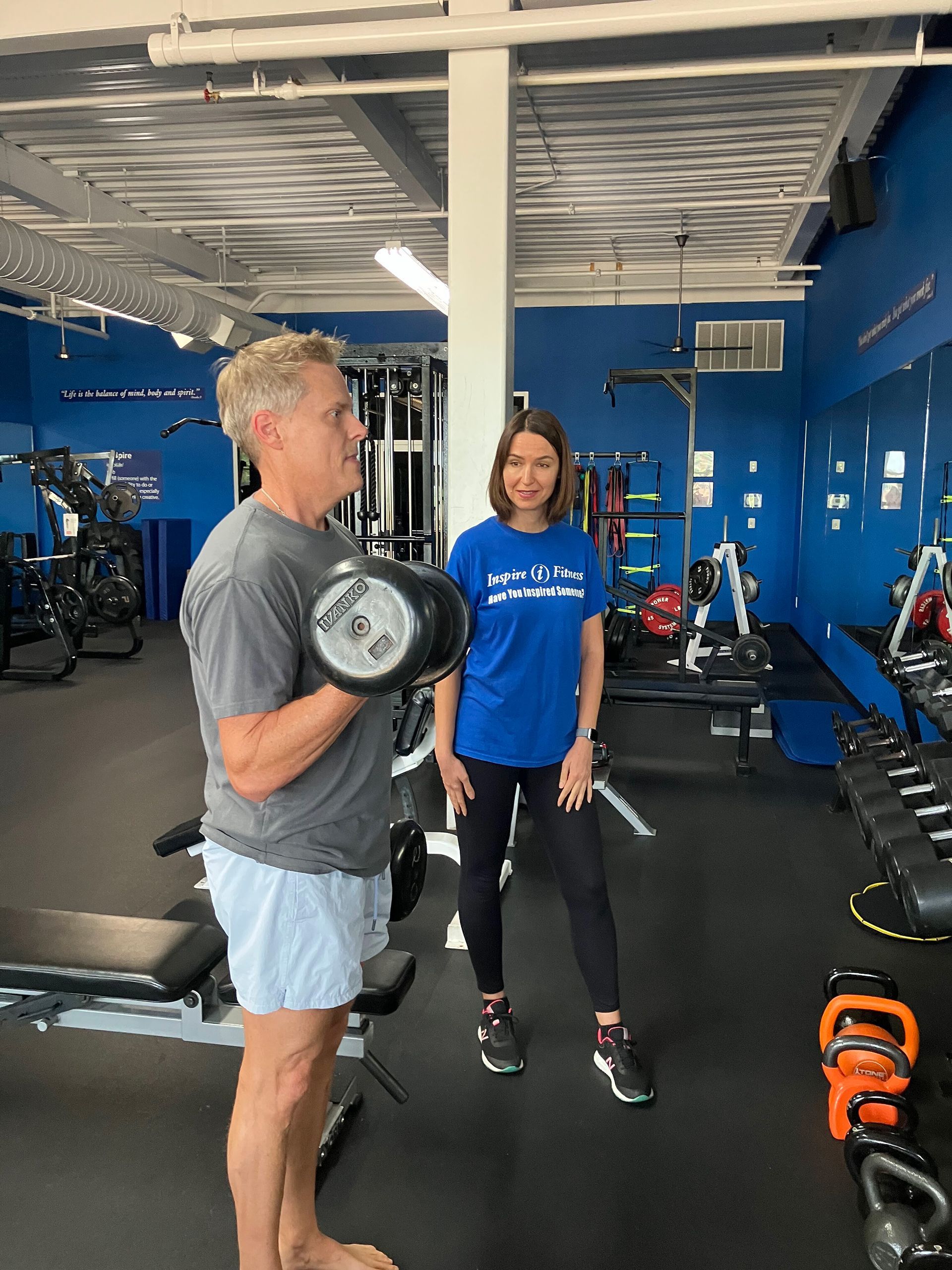 Two people in a blue gym, one holding a dumbbell while standing beside the other.