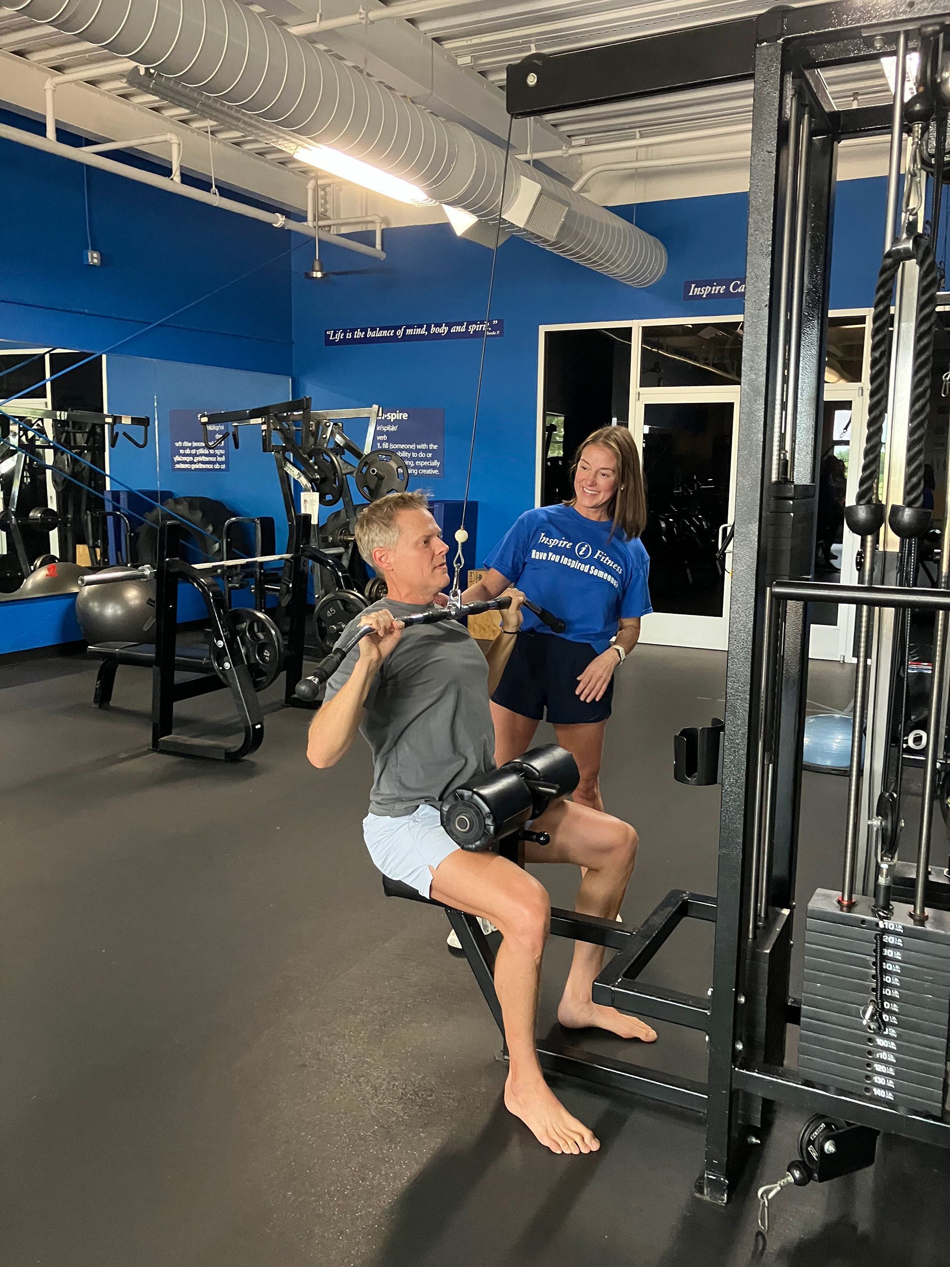 Two people at a gym using a cable machine, one seated and pulling the handle while the other stands nearby.
