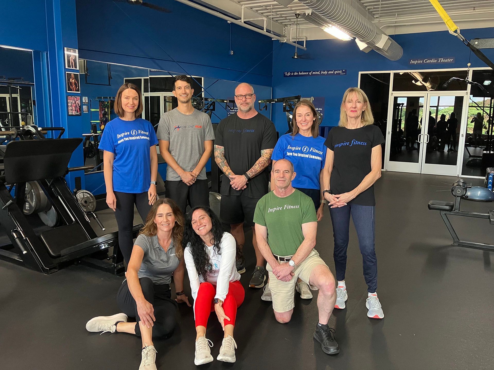 Group of seven people posing in a gym with exercise equipment and blue walls.