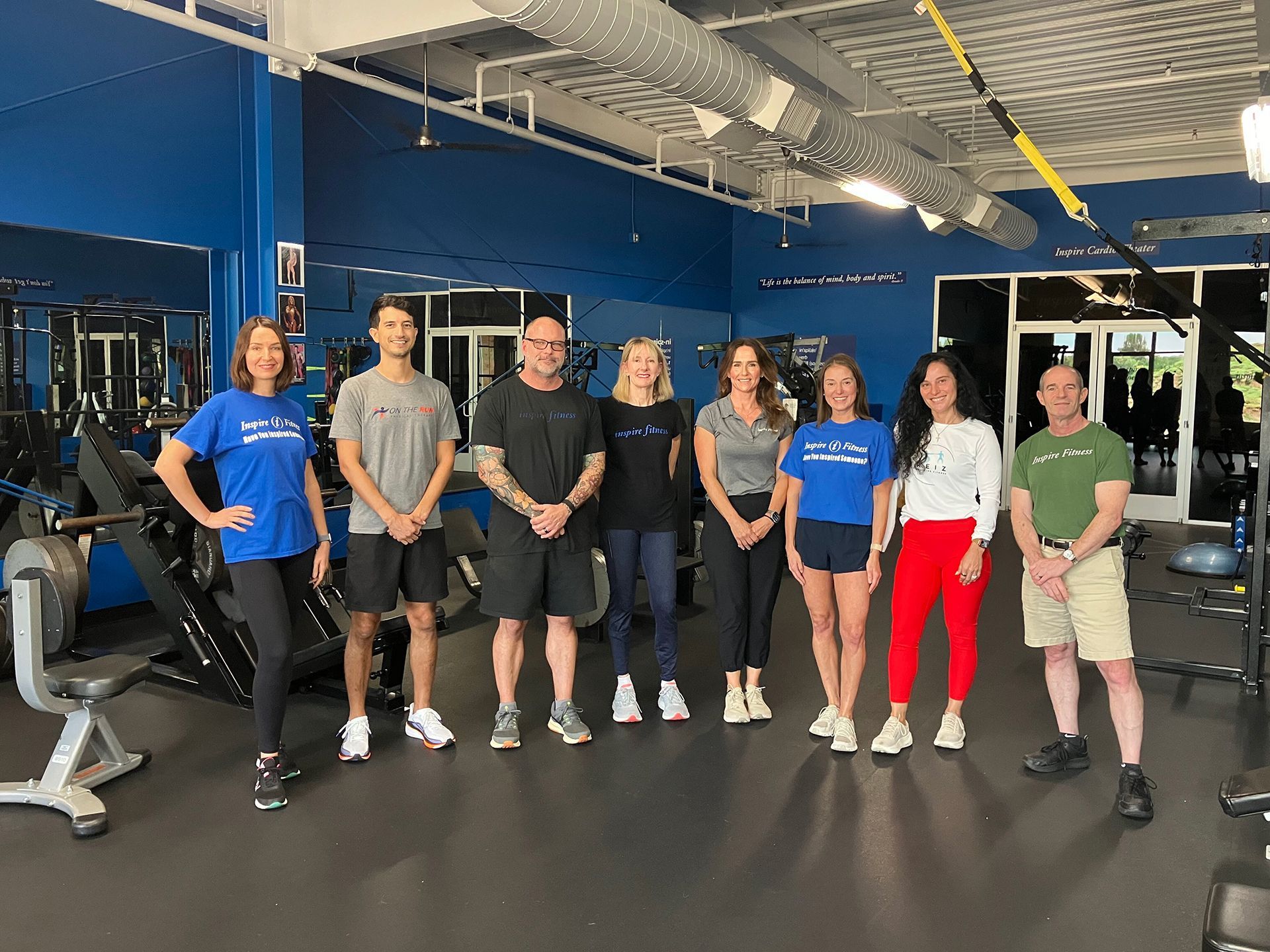 Group of seven people standing in a gym, posing in front of workout equipment and blue walls.