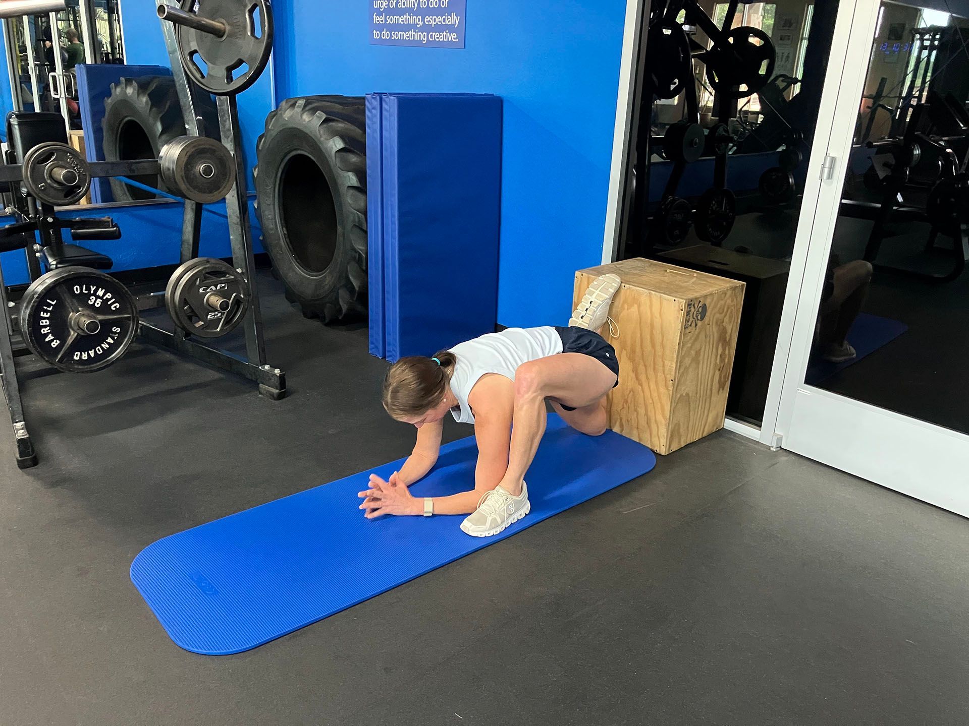 Person stretching on a blue mat in a gym, leaning forward with hands on the floor.
