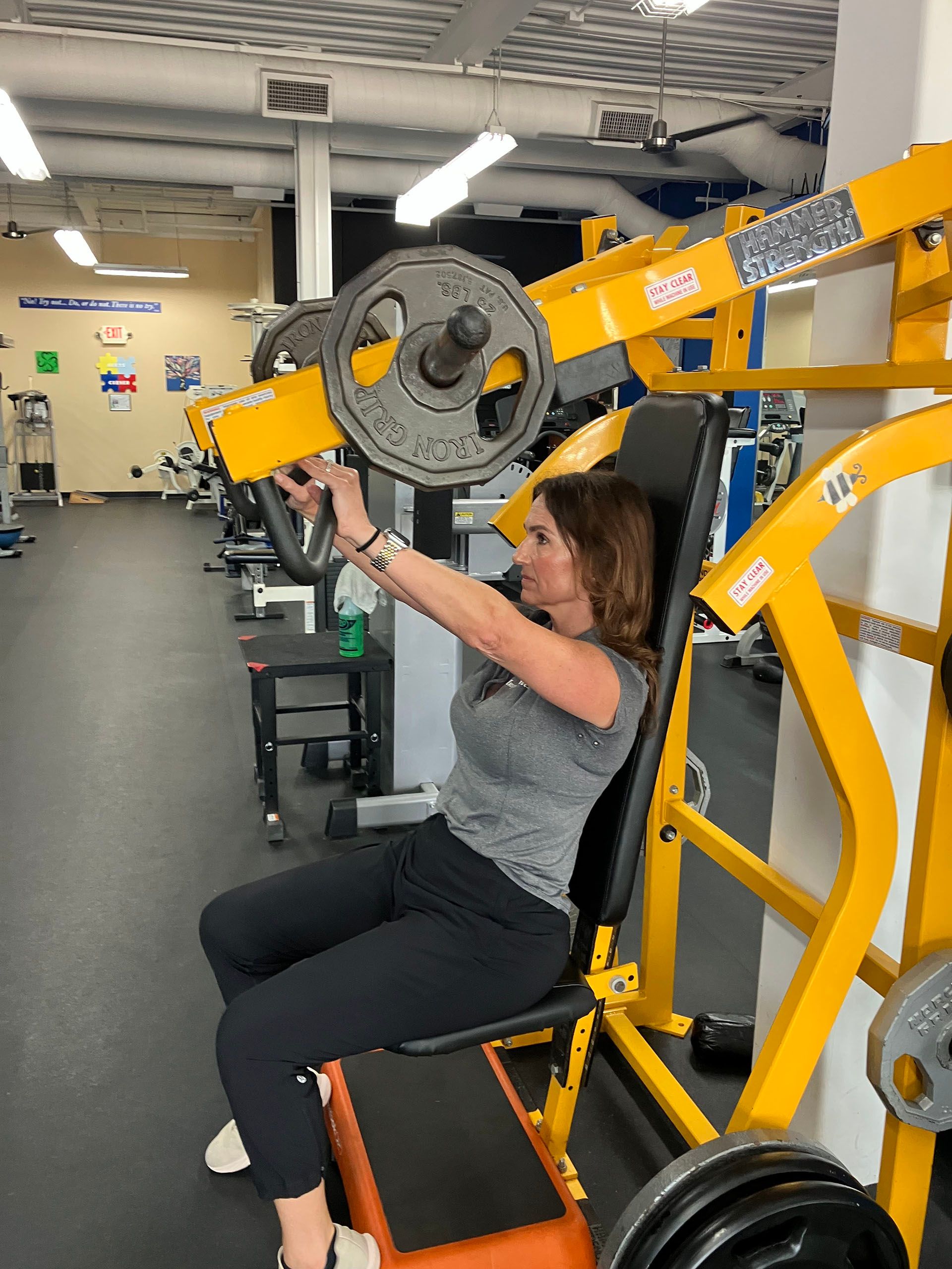 Person using a yellow gym machine in a fitness room, seated and holding the handles.