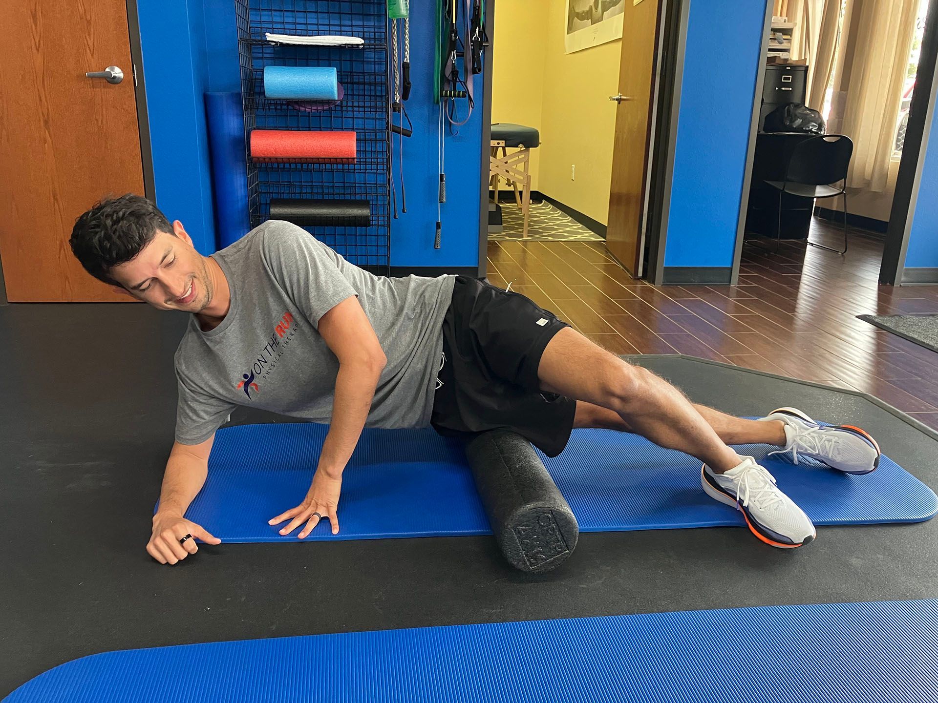 Person lying on a blue foam roller on a gym floor, stretching on a mat near exercise equipment.