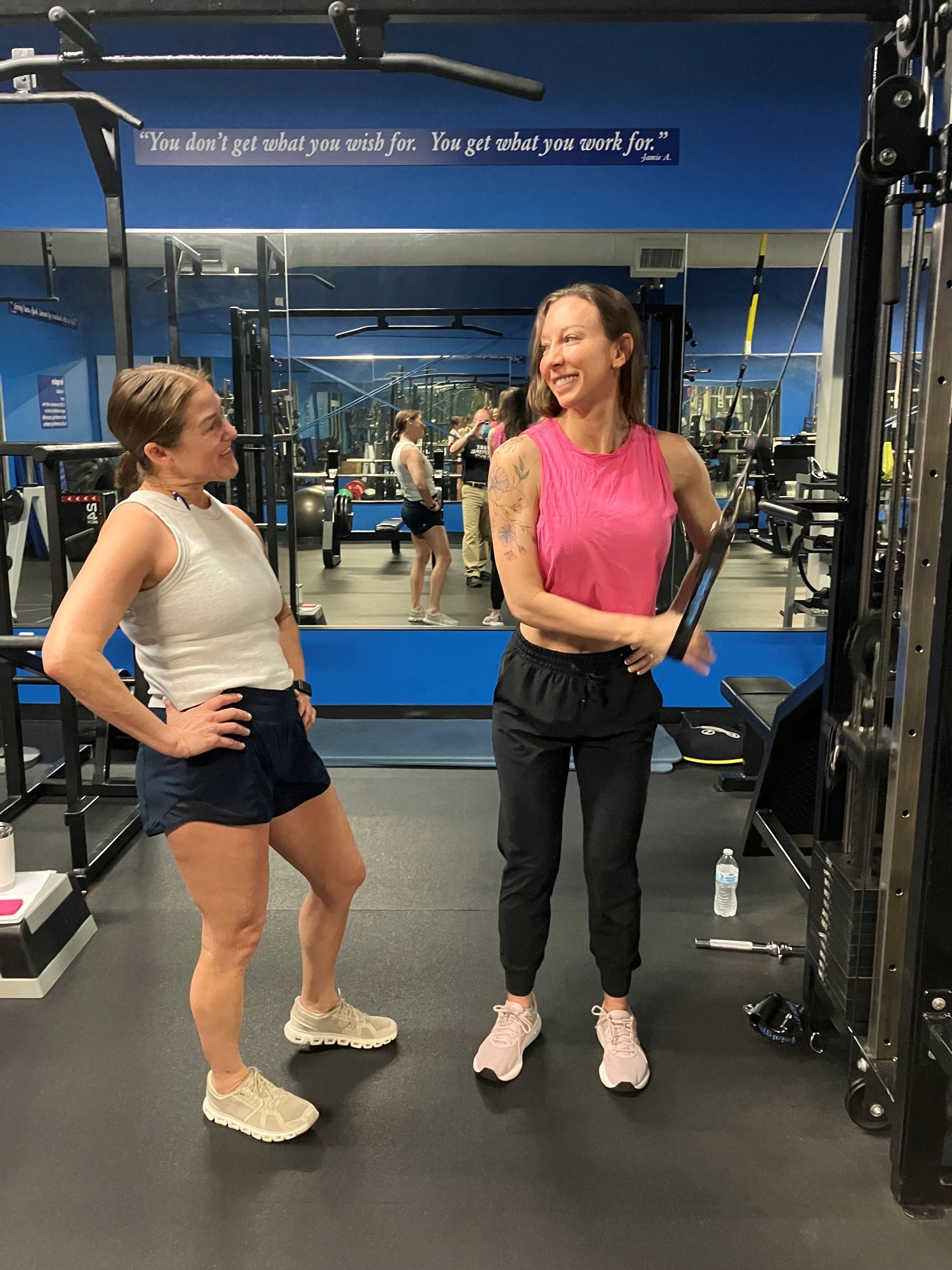 Two women in a gym, one smiling while holding a fitness handle near a cable machine; another woman stands nearby.