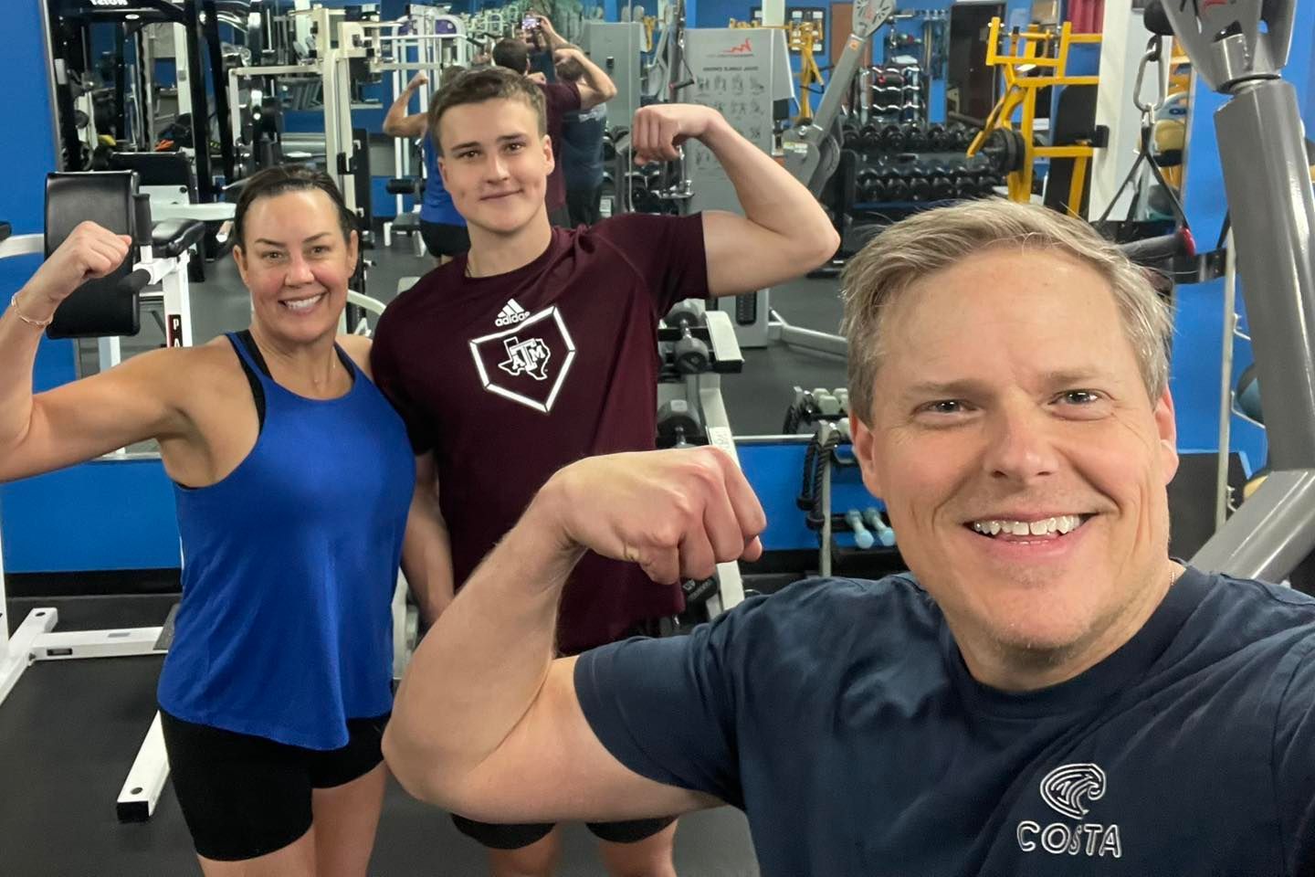 Three people flexing in a gym. A woman, and two men, all smiling, showing their muscles.
