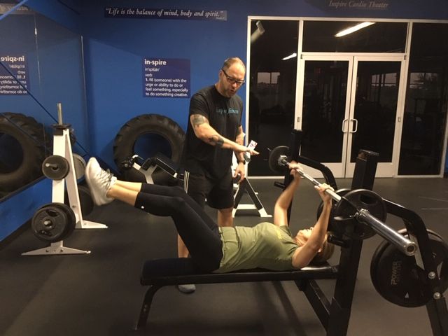 Woman on bench press, being coached. Gym setting with blue wall and equipment.