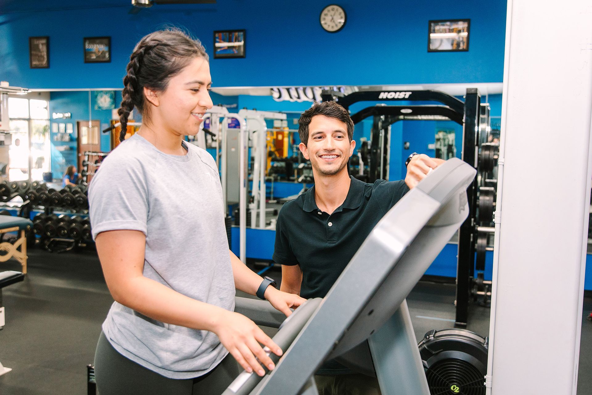 Woman on treadmill with trainer, smiling in gym. Blue walls, gym equipment in background.