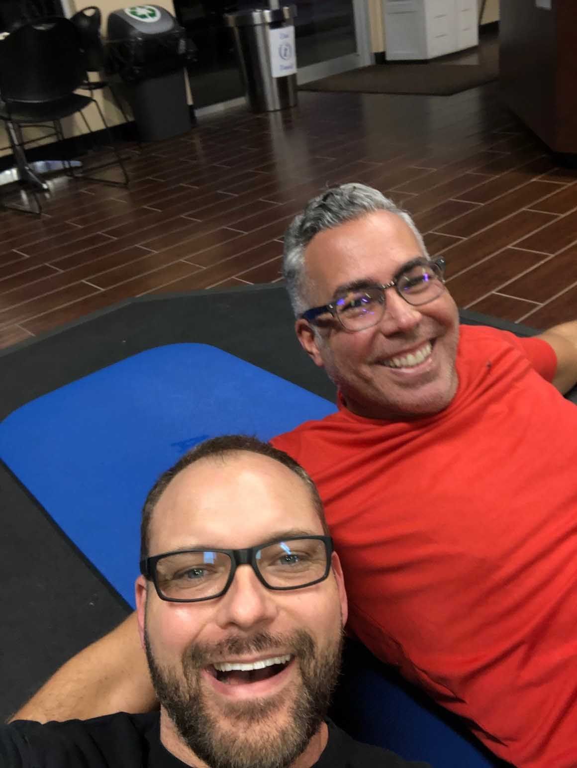 Two smiling men, one in red shirt, the other in black, taking a selfie on a blue mat in a room with wood flooring.