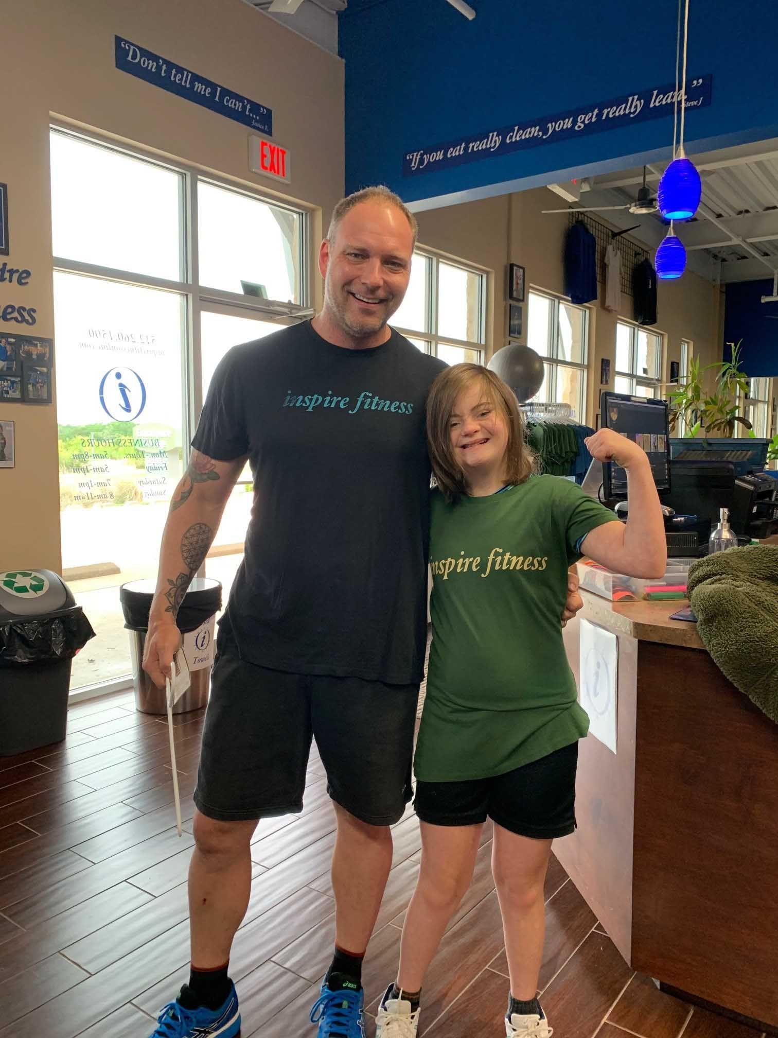 Man and girl posing at Inspire Fitness. Girl flexes arm, both wear green shirts; setting is a gym.