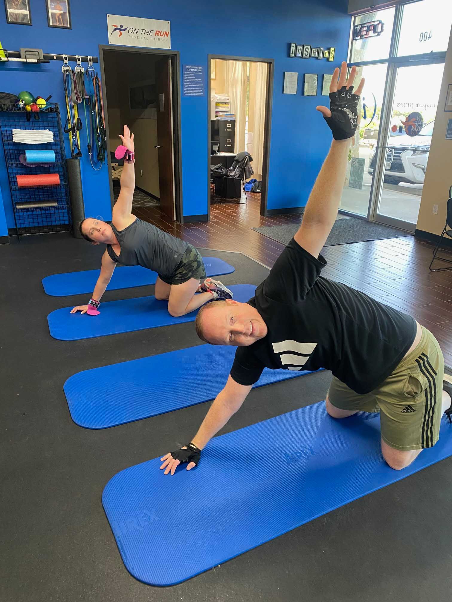 Two people exercise on blue mats in a gym, doing a torso twist.