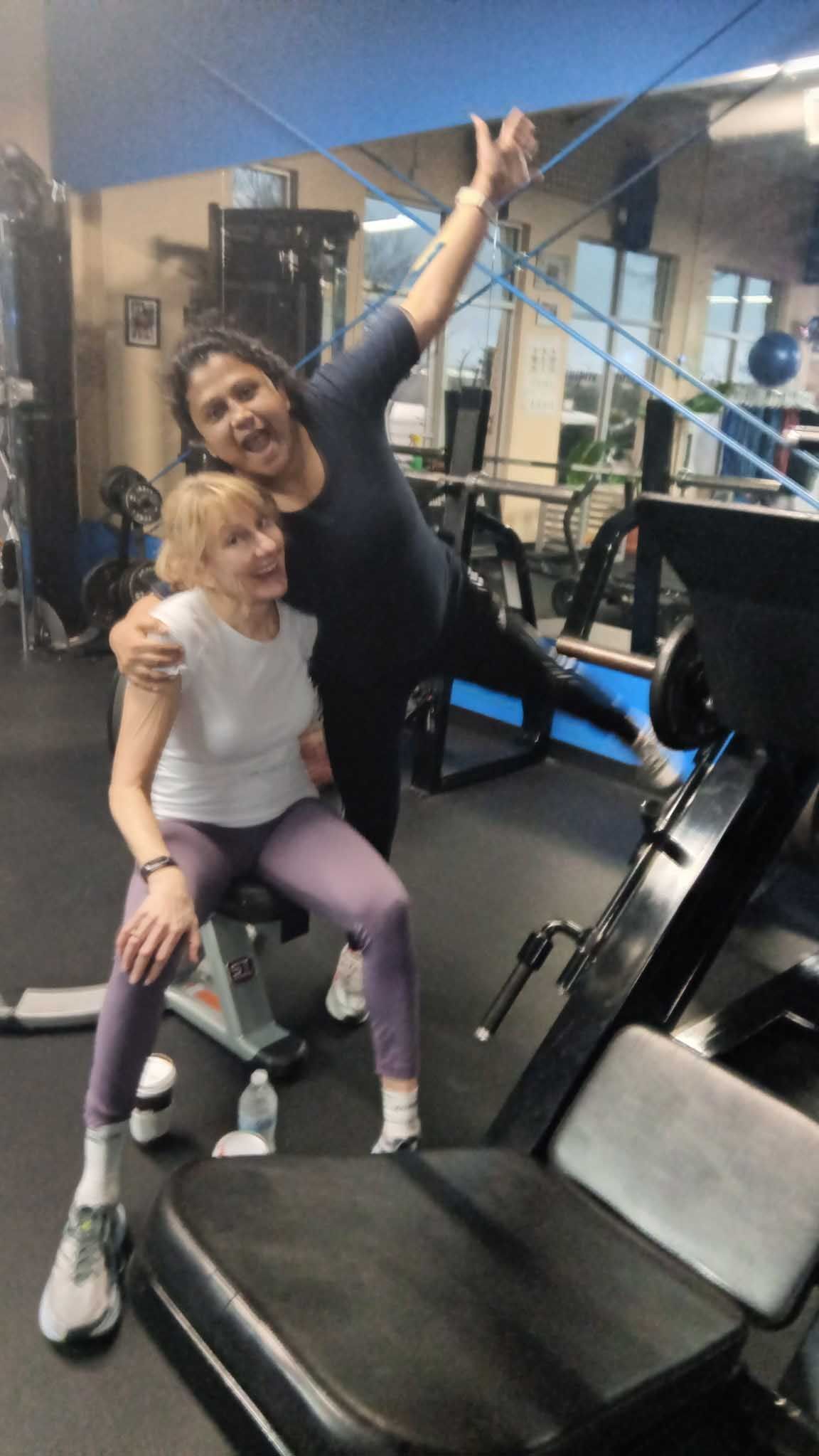 Woman in gym with trainer, poses with leg raised. Trainer smiles, arm around woman.