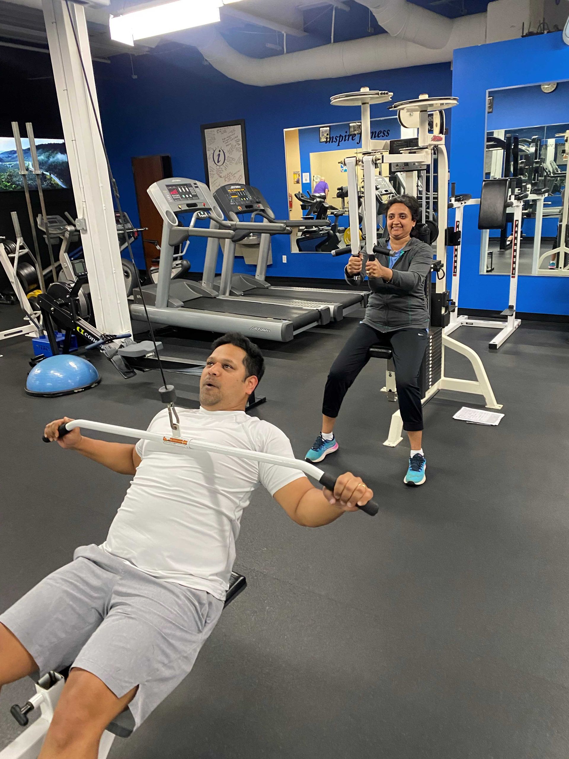 Two people exercising in a gym. Man rows a machine, woman does standing arm exercises. Blue walls.