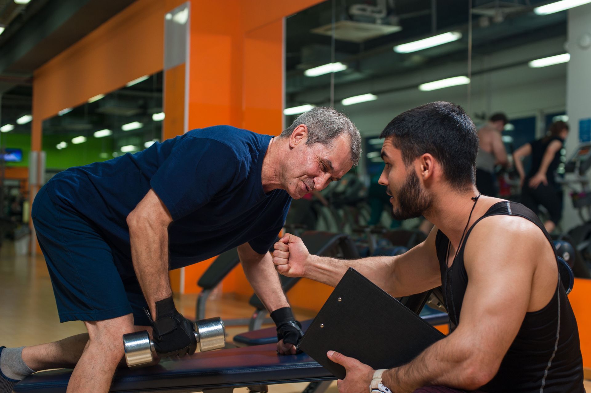 Older man lifting weights with a trainer at a gym. Trainer observes, holding a clipboard.