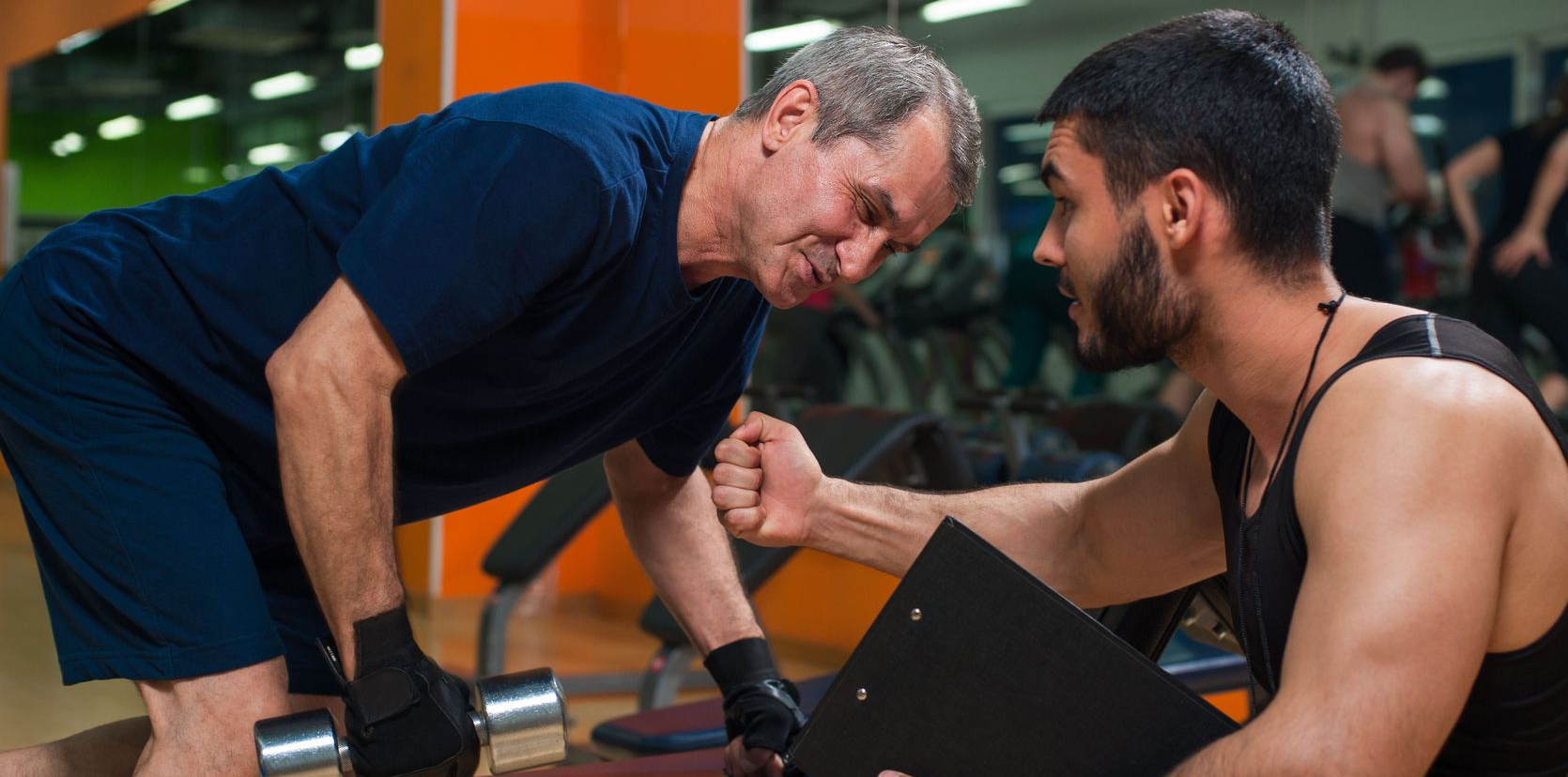 Older man lifting weights with a trainer at a gym. Trainer observes, holding a clipboard.