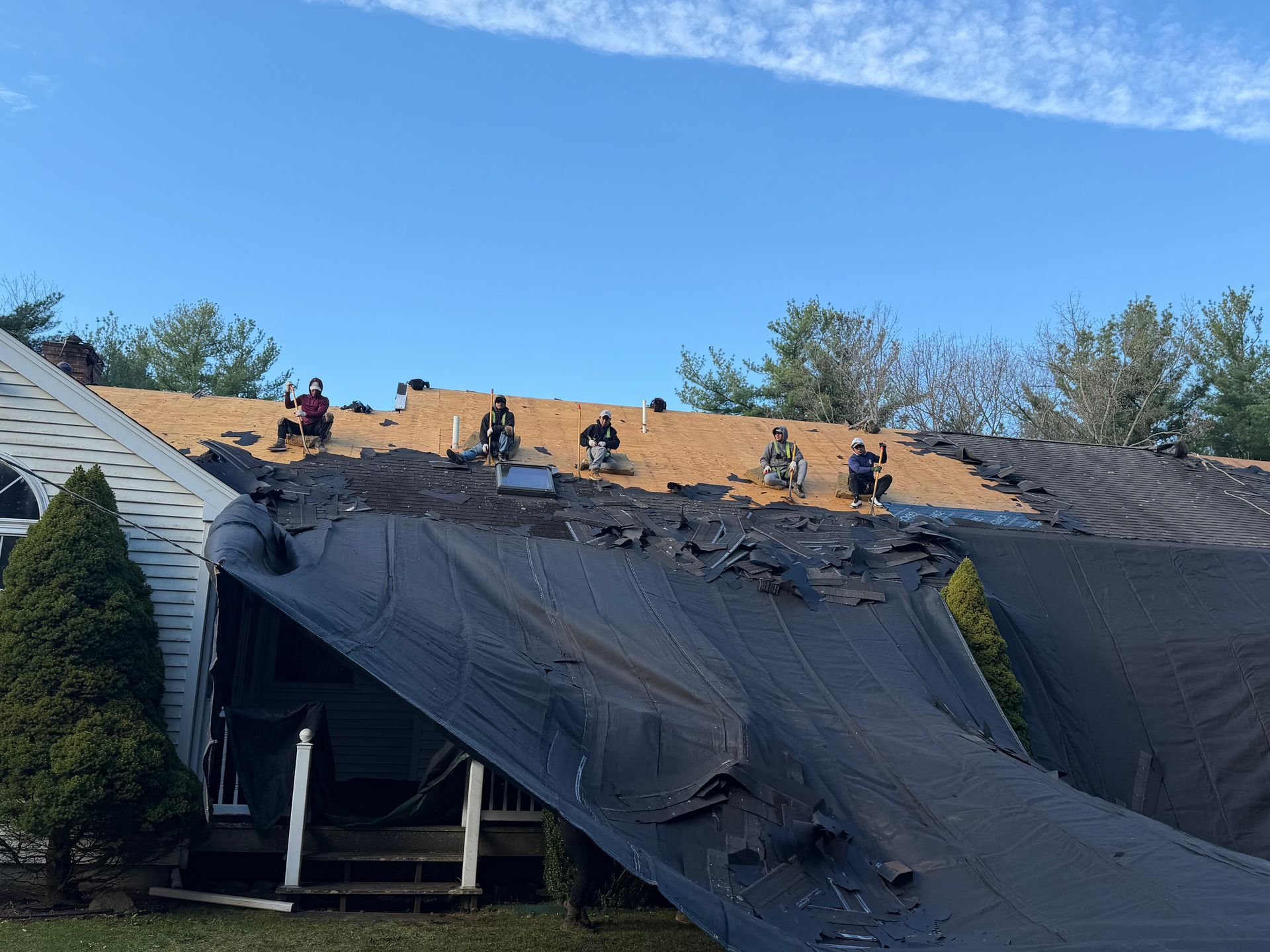 Roofers working on a house roof with black tarp and plywood under a blue sky
