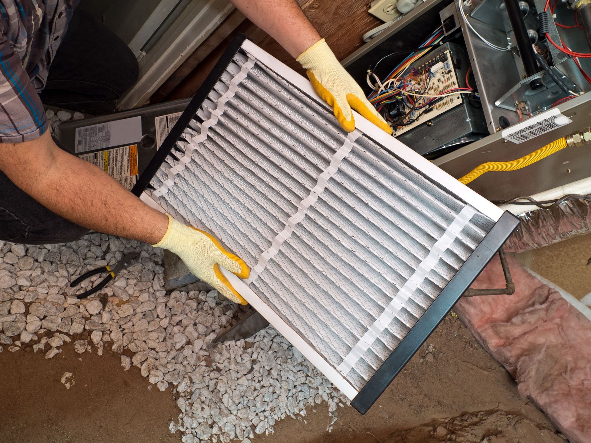 A person in yellow gloves holds a clean, rectangular air filter in front of an open HVAC furnace unit.
