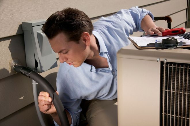 A technician in a light blue shirt inspects the exterior electrical box and air conditioning unit of a home.