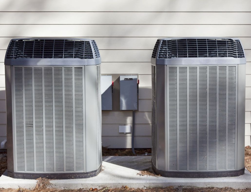 Two outdoor HVAC condenser units sit side-by-side on a concrete pad against a beige horizontal-siding wall.