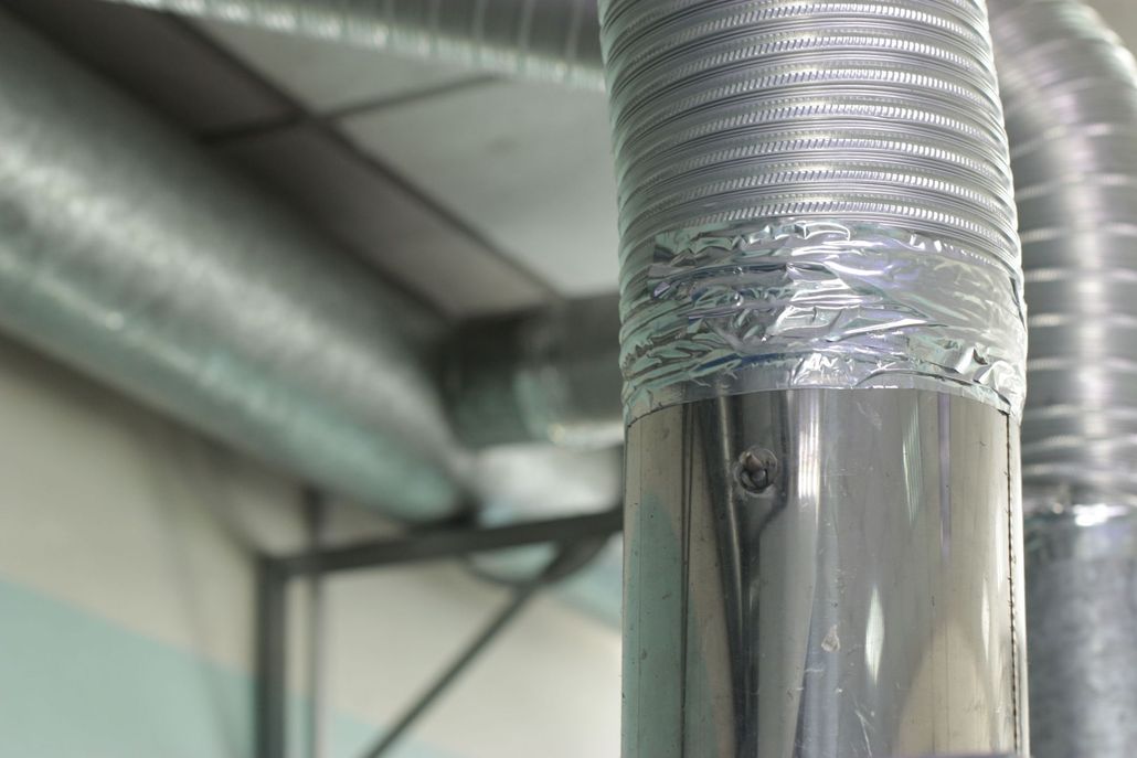 A close-up of flexible metallic ducting connected to a smooth metal pipe with silver tape in an industrial ceiling setting.