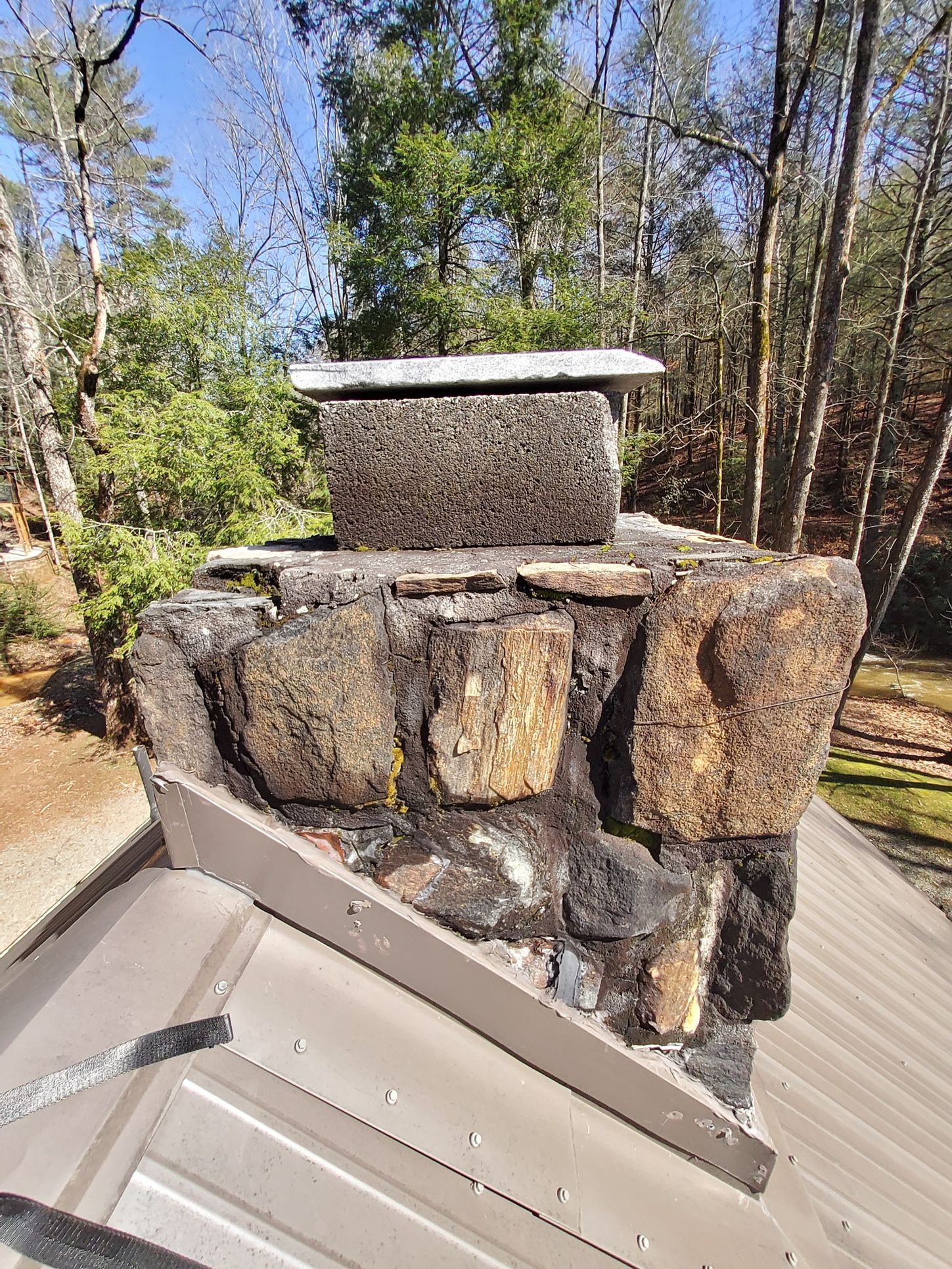 A stone chimney on top of a metal roof with trees in the background.