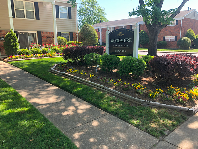 Apartment complex sign with manicured landscaping, including flowers and shrubs, and a sidewalk.