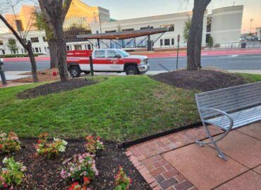 Red and white truck parked near a grassy hill with flowers and a bench in front of a building.