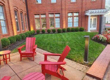 Red Adirondack chairs on a patio beside a lawn with a brick building in the background.