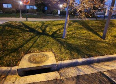 Concrete curb with open drain, grassy area, and tree shadows at night.