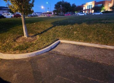 Asphalt parking lot with grass border and small tree; dusk setting with street lights and buildings in the background.