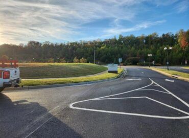 A paved road with white lane markings leads into a residential area, with a grassy roundabout and trees in the background.