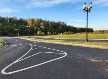 Asphalt road with white painted turning lane markers. Street lights line the road; background has green field and trees under blue sky.