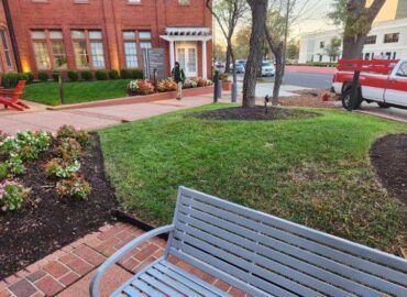 Lawn with a silver bench in the foreground, red brick walkway, flower beds, and a red brick building. A person walks by.