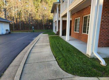 Sidewalk and grass border a building with a red brick facade. Sunny outdoor setting.