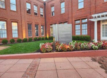 Brick building with a sign in front, surrounded by flowers, grass, and bushes. Paved walkway in foreground.