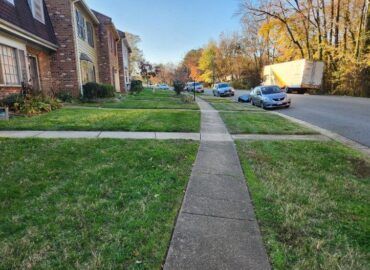 A paved sidewalk extends through a grassy area in front of townhouses. Cars parked on the street. Trees in background.