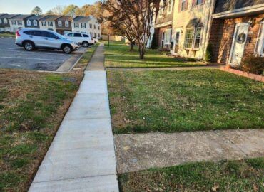 Sidewalk next to grass and townhouses with parked cars.