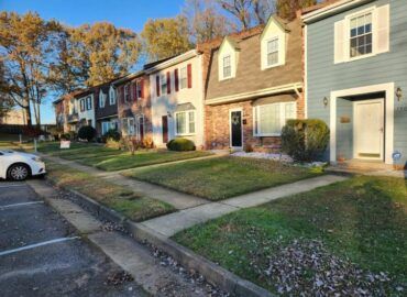 Row of townhouses with varying colors and architectural styles, along a sidewalk and lawn.